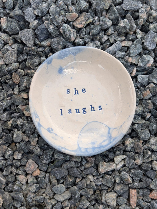 She Laughs round trinket tray in blue bubble glaze – a handmade ceramic dish about 10.5cm wide, hand-stamped with the words “she laughs”, shown from above on grey stone chippings