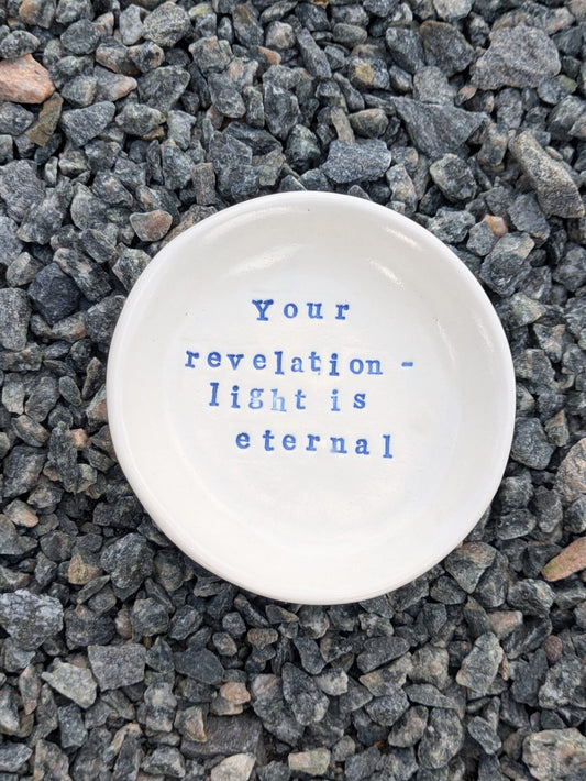 Your Revelation - Light Is Eternal round trinket tray – a handmade ceramic dish about 10.5cm wide, hand-stamped with the words “your revelation - light is eternal”, shown from above on grey stone chippings