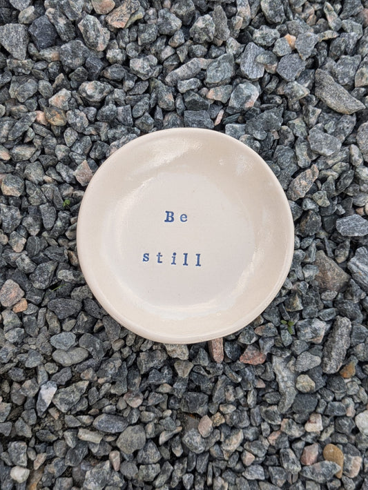 Be Still round trinket tray – a handmade ceramic dish about 10cm wide, hand-stamped with the words “be still”, shown from above on grey stone chippings