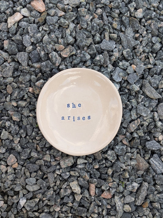 She Arises round trinket tray – a handmade ceramic dish about 10cm wide, hand-stamped with the words “she arises”, shown from above on grey stone chippings