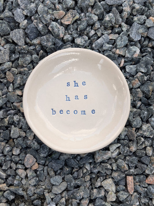 She Has Become round trinket tray – a handmade ceramic dish about 10cm wide, hand-stamped with the words “she has become”, shown from above on grey stone chippings