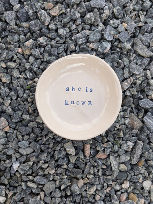 She Is Known round trinket tray – a handmade ceramic dish about 10cm wide, hand-stamped with the words “she is known”, shown from above on grey stone chippings