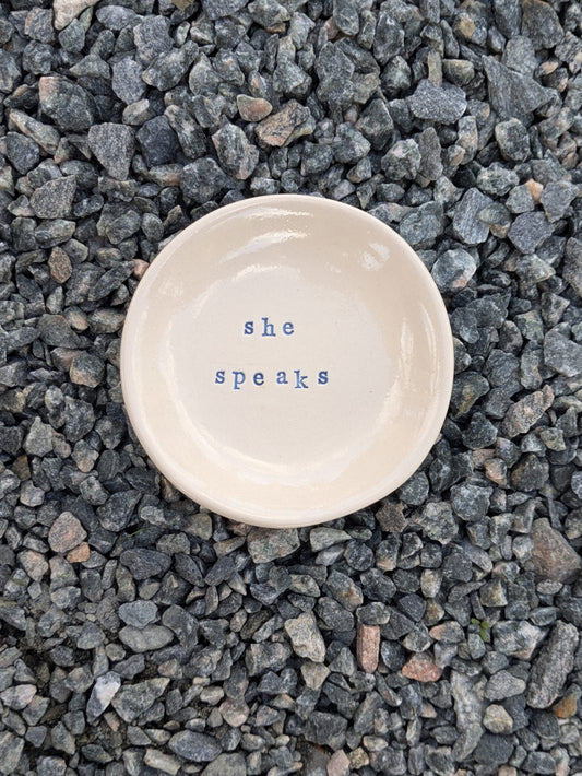 She Speaks round trinket tray – a handmade ceramic dish about 10.5cm wide, hand-stamped with the words “she speaks”, shown from above on grey stone chippings