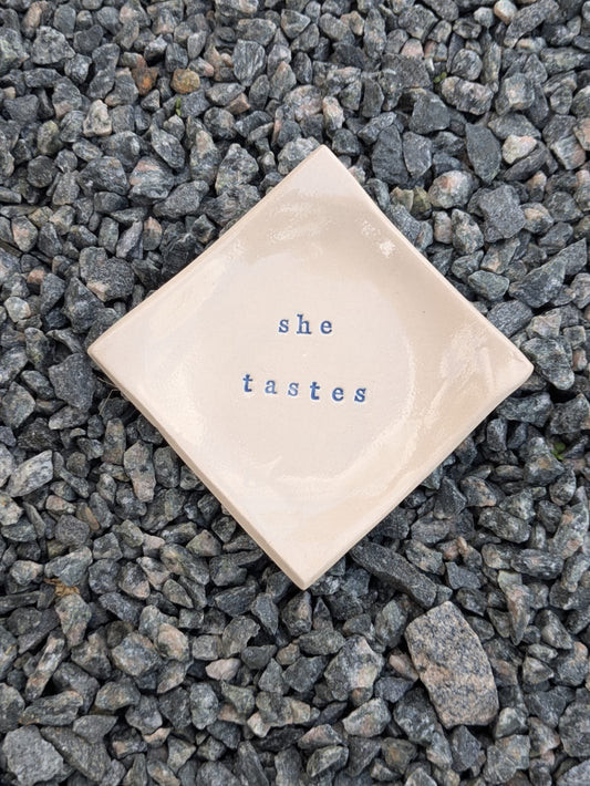 She Tastes square trinket tray – a handmade ceramic dish about 9.5cm square, hand-stamped with the words “she tastes”, shown from above on grey stone chippings