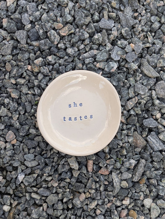 She Tastes round trinket tray – a handmade ceramic dish about 10.5cm wide, hand-stamped with the words “she tastes”, shown from above on grey stone chippings