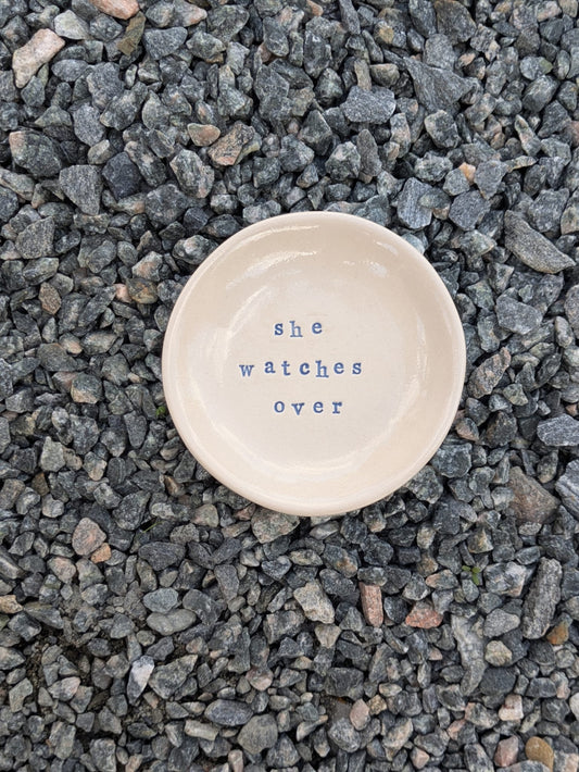 She Watches Over round trinket tray – a handmade ceramic dish about 10.5cm wide, hand-stamped with the words “she watches over”, shown from above on grey stone chippings