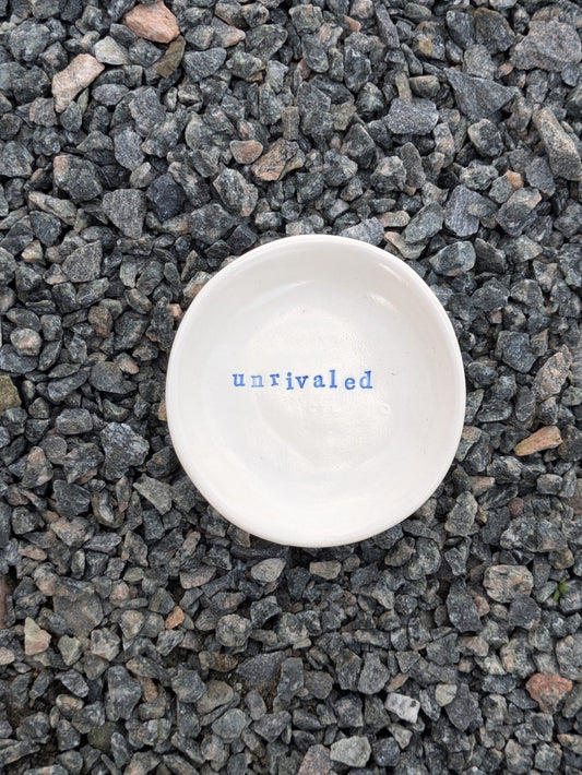 Unrivaled round trinket tray – a handmade ceramic dish about 10.5cm wide, hand-stamped with the words “unrivaled”, shown from above on grey stone chippings