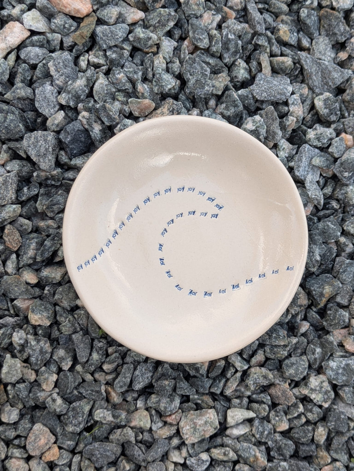Wave of Joy round trinket tray – a handmade ceramic dish about 10.5cm wide, decorated with a hand-stamped wave pattern, shown from above on grey stone chippings